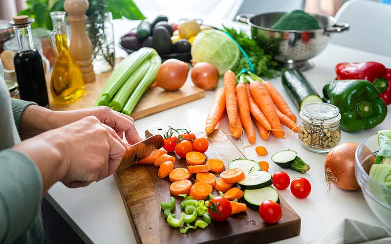 Eine Frau, von der nur die Hände zu sehen sind, schneidet in einer Küche auf einem Schneidebrett verschiedene Gemüsesorten: Karotten, Tomaten, Sellerie und Zucchini. Weiteres Gemüse liegt auf der Arbeitsfläche.