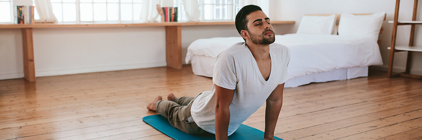 Ein junger Mann macht zuhause Yoga-Übungen. Mit den Händen stützt er sich auf einer Gymnastikmatte ab und hebt den Oberkörper. Der Kopf ist leicht nach hinten geneigt, die Augen sind geschlossen, Seine Beine sind auf der Matte abgelegt. 