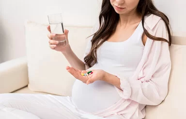 Eine schwangere Frau mit langen braunen Haaren, deren Kopf nicht ganz zu sehen ist, sitzt auf einem weißen Sofa und hält in der einen Hand verschiedene bunte Tabletten und Kapseln und in der anderen Hand ein Glas mit Wasser.