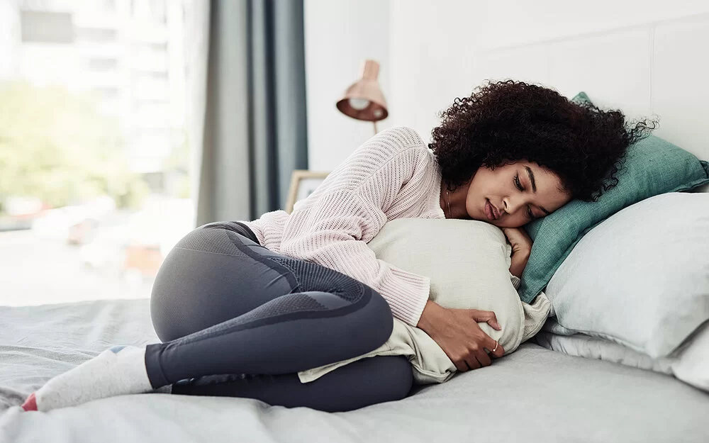 Eine Frau liegt mit angezogenen Beinen und einem Kissen, das sie sich vor den Bauch hält, auf einem Bett.