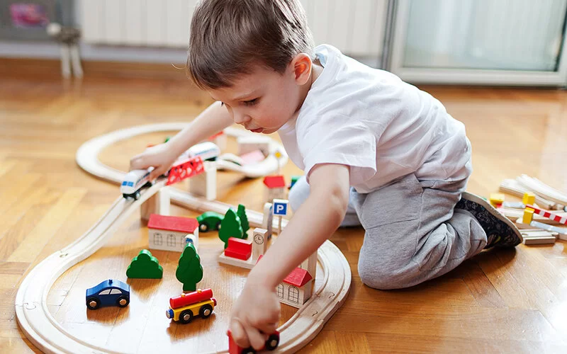 Ein etwa dreijähriger Junge spielt mit einer Holzeisenbahn in einem Zimmer mit Parkettfußboden.