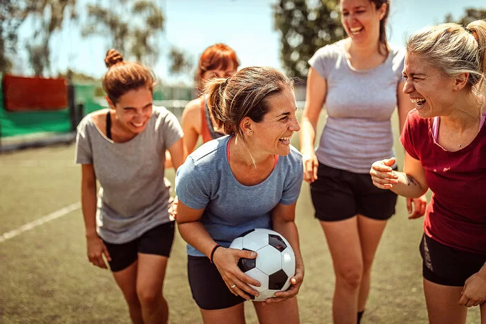 Eine Gruppe von Frauen in Sportkleidung steht lachend auf einem Fußballfeld. Die Frau in der Mitte hält einen Fußball. 