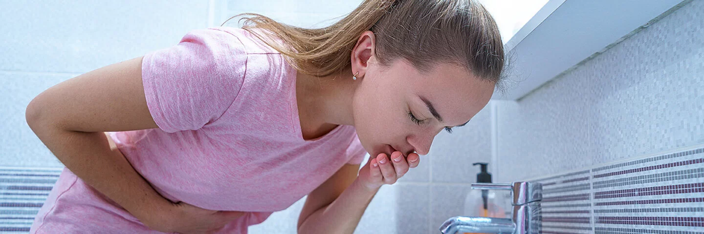 Eine junge Frau im Schlafanzug, die langen blonden Haare zum Zopf gebunden, lehnt sich im Badezimmer über ein Waschbecken und fasst sich mit der rechten Hand an den Mund. Ihr ist übel.