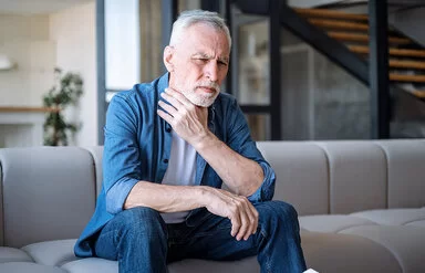 Ein älterer Mann mit kurzem weißen Vollbart sitzt auf einem Sofa und fasst sich mit einer Hand an den Hals. Sein Gesicht drückt Schmerz oder Unbehagen aus.