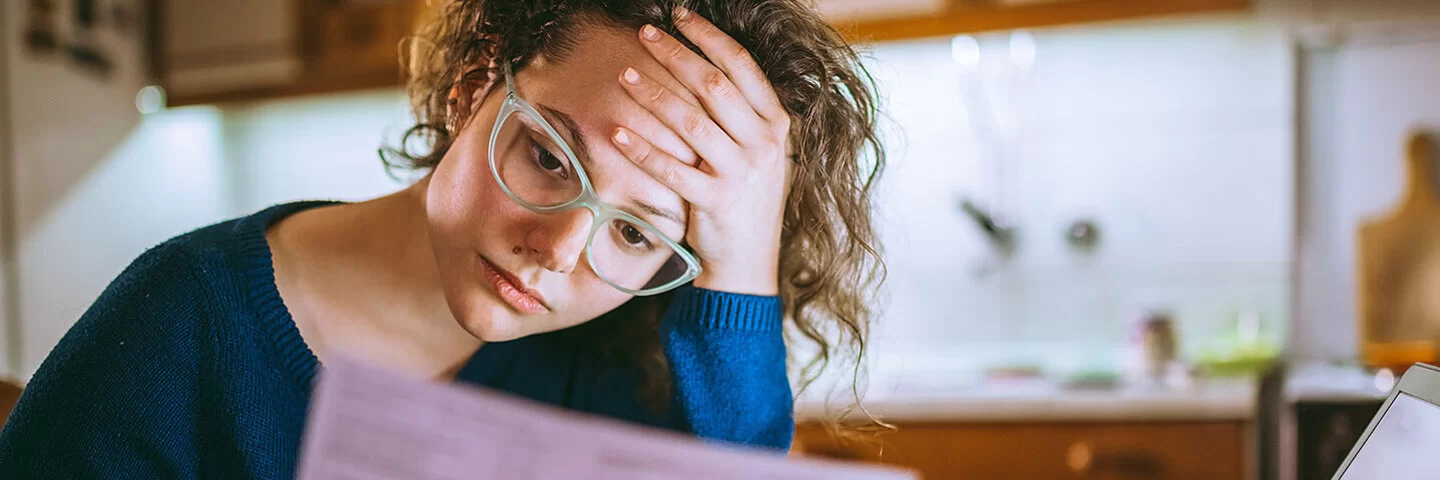 Junge Frau, die mit Rechnungen in der Hand vor ihrem aufgeklappten Laptop am Küchentisch sitzt und sich gestresst mit der linken Hand an die Stirn fasst.