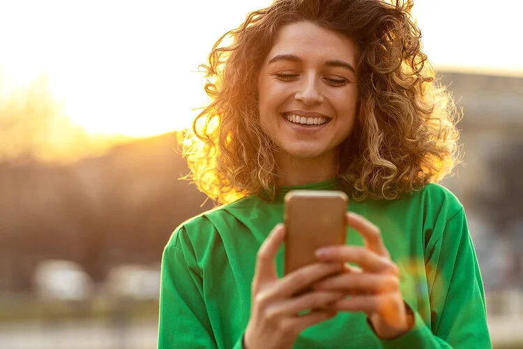 Eine Frau mit schulterlangem lockigen Haar blickt fröhlich lachend auf ihr Smartphone, im Hintergrund sieht man die Sonne untergehen.