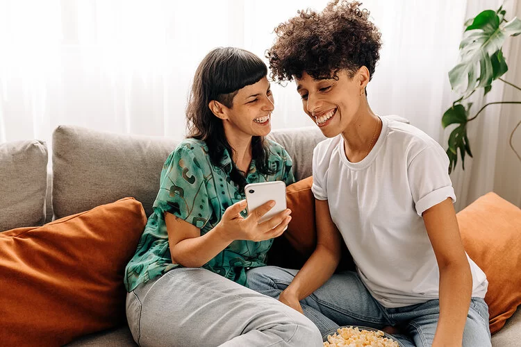 Zwei junge Frauen sitzen eng beieinander auf einem Sofa, eine der beiden zeigt der anderen etwas auf ihrem Smartphine. Beide lachen und wirken fröhlich.