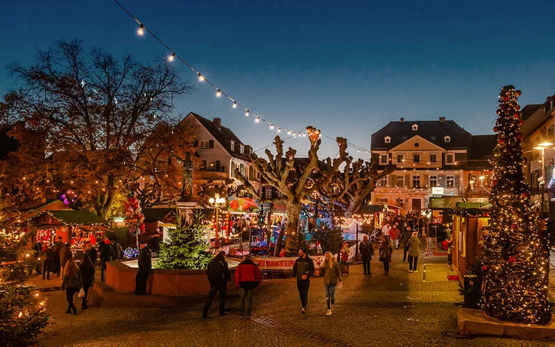 Abendliche Szene, Marktplatz mit Weihnachtsmarkt, bunte Beleuchtung und Passanten.