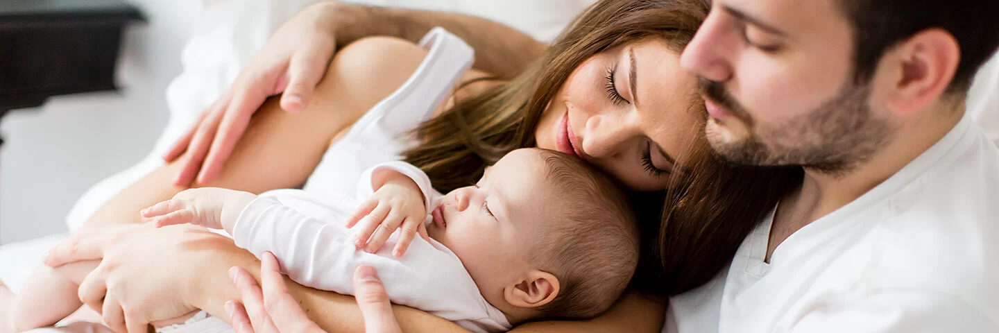 Ein Mann mit kurzem braunem Bart und weißem T-Shirt sitzt auf einem Bett und umarmt eine Frau mit langen braunen Haaren. Die Frau hält ein kleines schlafendes Baby im Arm.