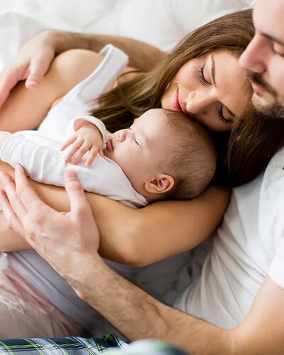 Ein Mann mit kurzem braunem Bart und weißem T-Shirt sitzt auf einem Bett und umarmt eine Frau mit langen braunen Haaren. Die Frau hält ein kleines schlafendes Baby im Arm.