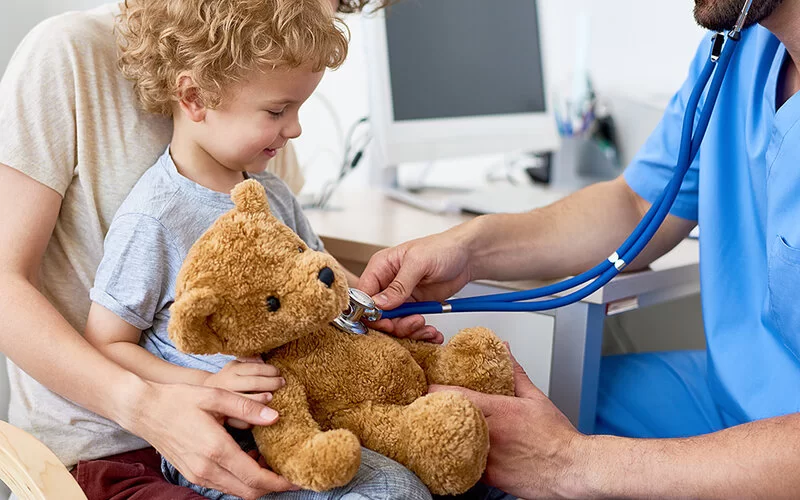 Eine Mutter sitzt mit ihrem kleinen Sohn im Behandlungsraum eines Kinderarztes. Zur Auflockerung der Situation hört der Arzt mit einem Stethoskop den Teddy ab, der auf dem Schoß des Kindes liegt.