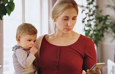 Eine etwa 30-jährige Frau mit blonden zusammengebundenen Haaren, weinrotem T-Shirt und weißem Cardigan steht mit ihrem Smartphone in der Hand im Wohnzimmer und hält ihr Baby im anderen Arm.