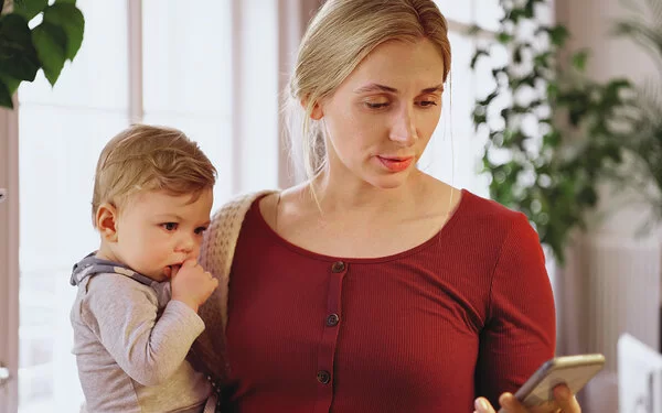 Eine etwa 30-jährige Frau mit blonden zusammengebundenen Haaren, weinrotem T-Shirt und weißem Cardigan steht mit ihrem Smartphone in der Hand im Wohnzimmer und hält ihr Baby im anderen Arm.