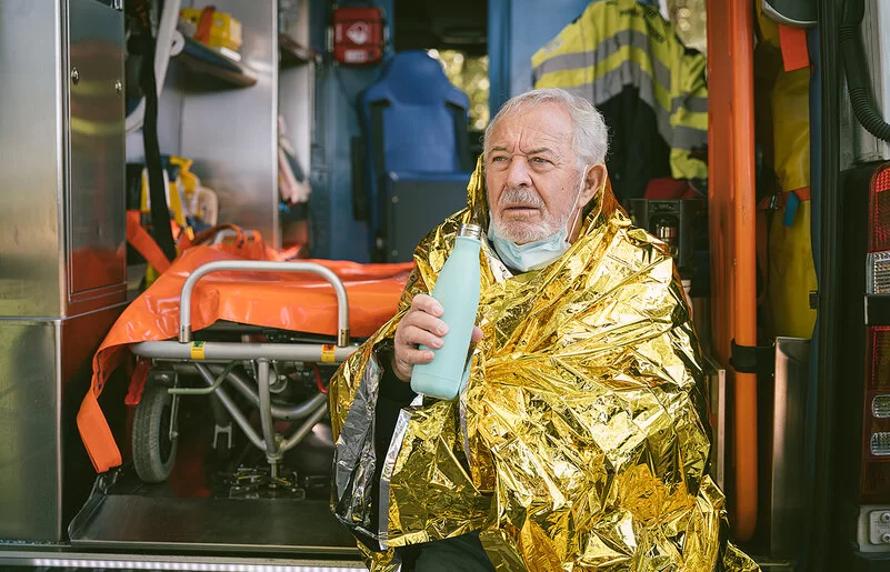 Ein älterer Mann sitzt in eine goldfarbene Wärmedecke gehüllt vor einem Krankenwagen und hält ein Heißgetränk in der Hand.