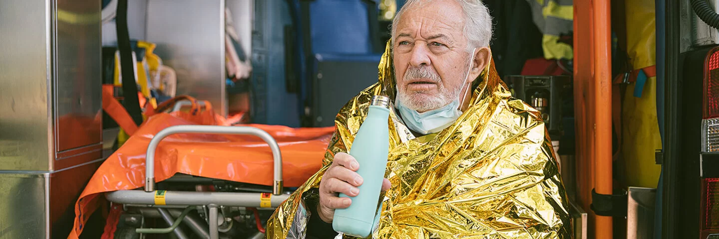 Ein älterer Mann sitzt in eine goldfarbene Wärmedecke gehüllt vor einem Krankenwagen und hält ein Heißgetränk in der Hand.