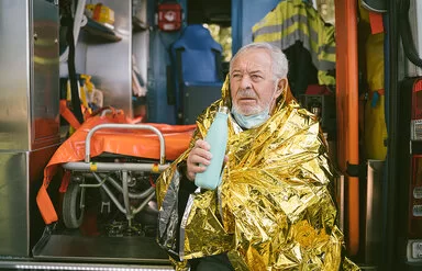 Ein älterer Mann sitzt in eine goldfarbene Wärmedecke gehüllt vor einem Krankenwagen und hält ein Heißgetränk in der Hand.