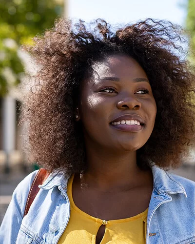 Ein junge Frau mit dunkler Haut und Locken steht in einem Park mit sattgrünen Bäumen. Ihr Blick ist zuversichtlich. Die Frau trägt ein gelbes Shirt und darüber eine hellblaue Jeans-Jacke.