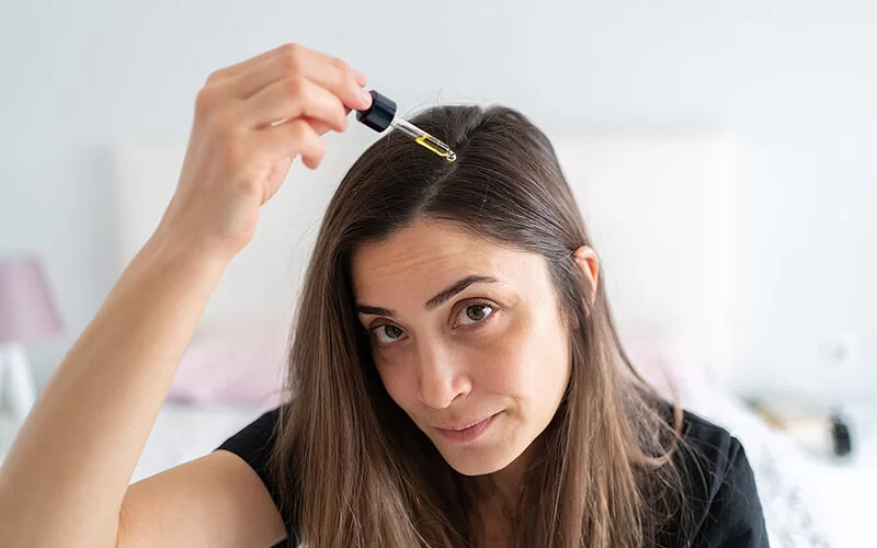 Eine Frau mit halblangen, glatten langen Haaren tropft sich mit einer Pipette eine gelbliche Flüssigkeit auf den Scheitel.