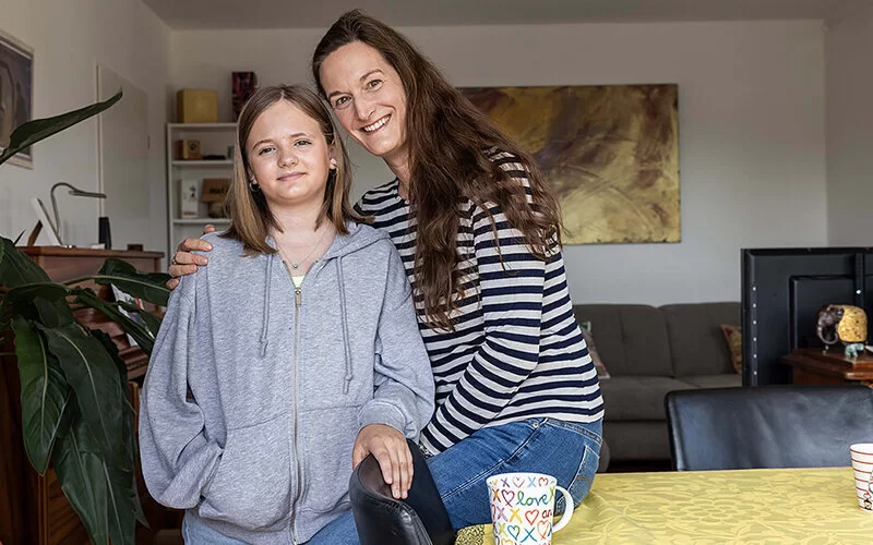 Eine Frau mit langen braunen Haaren im geringelten Shirt und ihre Tochter im Teenageralter im grauen Kapuzenpullover. Die Frau legt einen Arm um das Mädchen. Beide lächeln.