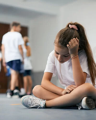 Eine Schülerin sitzt im Schneidersitz auf dem Boden der Turnhalle. Ihre Mitschüler und Mitschülerinnen stehen weit weg. Das Mädchen ist traurig und fühlt sich ausgeschlossen.