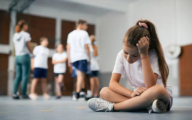 Eine Schülerin sitzt im Schneidersitz auf dem Boden der Turnhalle. Ihre Mitschüler und Mitschülerinnen stehen weit weg. Das Mädchen ist traurig und fühlt sich ausgeschlossen.