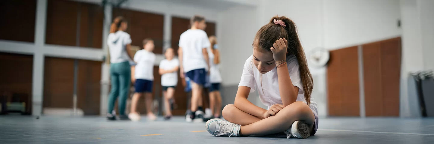 Eine Schülerin sitzt im Schneidersitz auf dem Boden der Turnhalle. Ihre Mitschüler und Mitschülerinnen stehen weit weg. Das Mädchen ist traurig und fühlt sich ausgeschlossen.