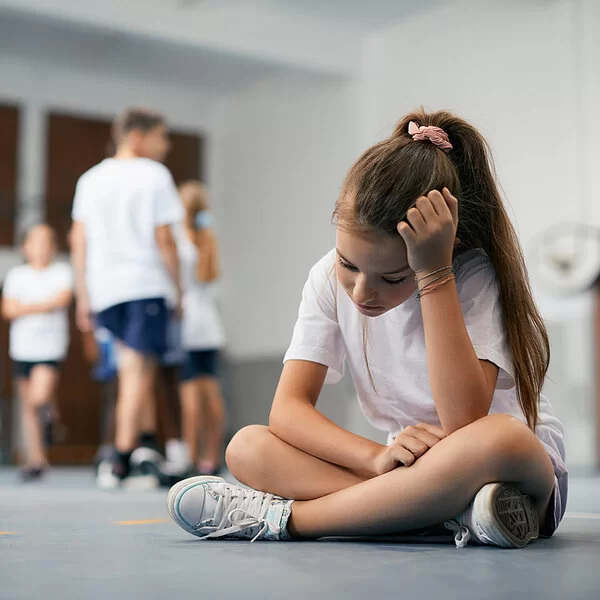 Eine Schülerin sitzt im Schneidersitz auf dem Boden der Turnhalle. Ihre Mitschüler und Mitschülerinnen stehen weit weg. Das Mädchen ist traurig und fühlt sich ausgeschlossen.