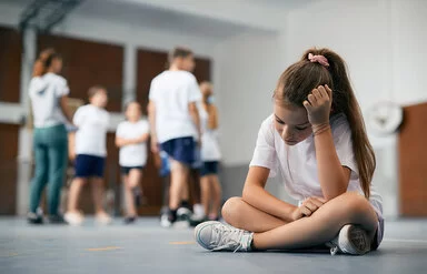 Eine Schülerin sitzt im Schneidersitz auf dem Boden der Turnhalle. Ihre Mitschüler und Mitschülerinnen stehen weit weg. Das Mädchen ist traurig und fühlt sich ausgeschlossen.