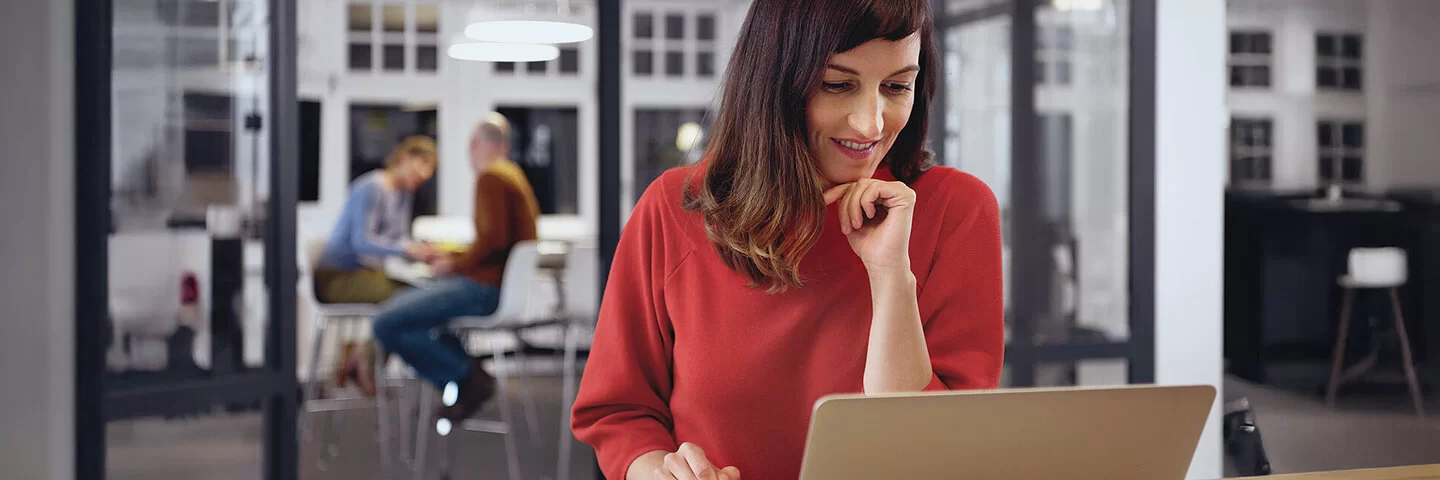 Eine Frau sitzt vor ihrem Laptop und informiert sich zum Zusatzbeitrag, im Hintergrund sitzen zwei Menschen in der Küche.