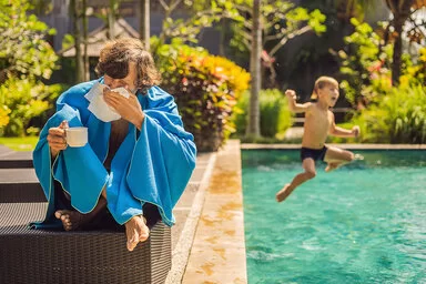 Ein Mann sitzt am Pool, in der einen Hand eine Tasse, in der anderen ein Taschentuch, mit dem er sich gerade die Nase putzt. Um seine Schultern hängt eine blaues Badelaken. Rechts neben ihm springt gerade ein kleiner Junge mit freudigem Gesicht in den Pool.