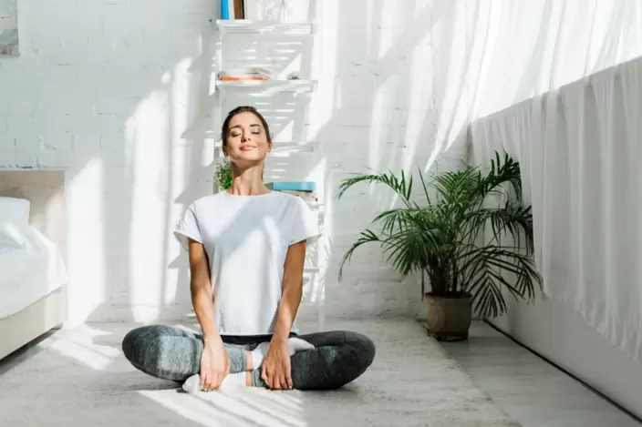 Eine junge Frau betreibt auf dem Boden ihres Schlafzimmers Yoga und hat die Augen geschlossen.