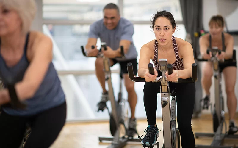 In einem Fitnessstudio trainiert eine Gruppe auf Fahrradergometern. In der Bildmitte trainiert eine junge Frau, drei weitere, ältere Personen sind unscharf abgebildet.