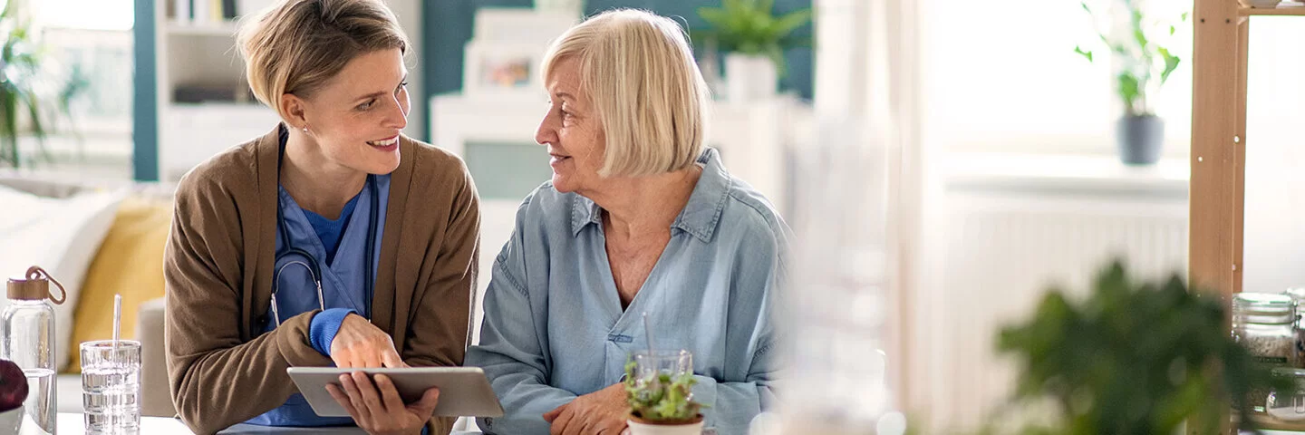 Eine freundliche Pflegekraft mit Stethoskop um den Hals sitzt mit einer älteren Frau an einem Tisch in einem hellen, gemütlichen Wohnzimmer. Die Pflegekraft zeigt der Seniorin etwas auf einem Tablet, während beide lächelnd miteinander sprechen. Auf dem Tisch stehen Pflanzen, Gläser und Unterlagen.