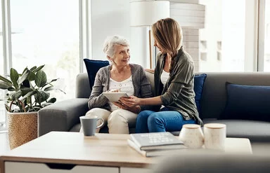 Eine freundliche junge Frau mit langem Haar sitzt mit einer älteren Frau auf einem Sofa in einem hellen, gemütlichen Wohnzimmer. Die junge Frau zeigt der Seniorin etwas auf einem Tablet, während beide lächelnd miteinander sprechen.