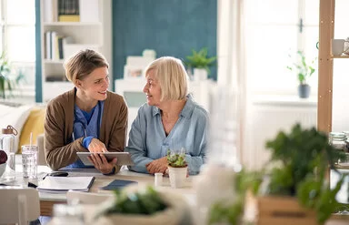 Eine freundliche Pflegekraft mit Stethoskop um den Hals sitzt mit einer älteren Frau an einem Tisch in einem hellen, gemütlichen Wohnzimmer. Die Pflegekraft zeigt der Seniorin etwas auf einem Tablet, während beide lächelnd miteinander sprechen. Auf dem Tisch stehen Pflanzen, Gläser und Unterlagen.