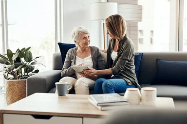 Eine freundliche junge Frau mit langem Haar sitzt mit einer älteren Frau auf einem Sofa in einem hellen, gemütlichen Wohnzimmer. Die junge Frau zeigt der Seniorin etwas auf einem Tablet, während beide lächelnd miteinander sprechen.