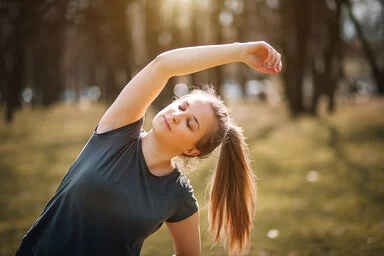 Das Bild zeigt eine Frau mit Pferdeschwanz und schwarzem T-Shirt, die ihren Körper draußen in der Natur seitlich streckt.