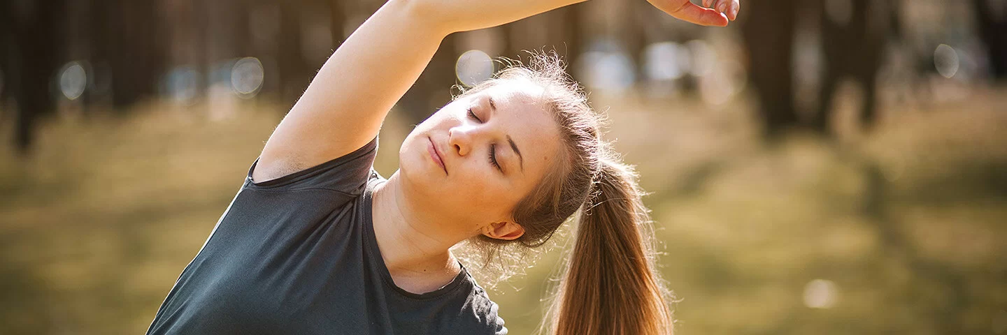 Das Bild zeigt eine Frau mit Pferdeschwanz und schwarzem T-Shirt, die ihren Körper draußen in der Natur seitlich streckt.