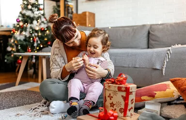 Ein kleiens Kind sitzt mit seiner Mutter auf dem Boden und öffnet mit ihr Weihnachtsgeschenke.