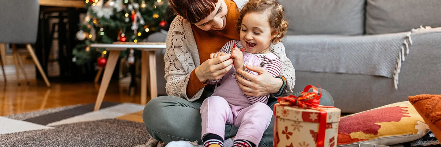 Ein kleiens Kind sitzt mit seiner Mutter auf dem Boden und öffnet mit ihr Weihnachtsgeschenke.