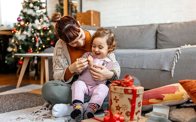 Ein kleiens Kind sitzt mit seiner Mutter auf dem Boden und öffnet mit ihr Weihnachtsgeschenke.