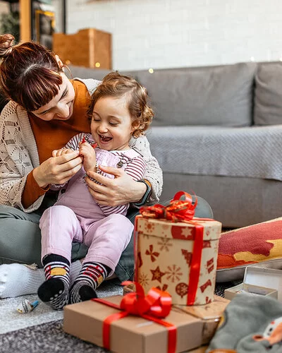 Ein kleiens Kind sitzt mit seiner Mutter auf dem Boden und öffnet mit ihr Weihnachtsgeschenke.