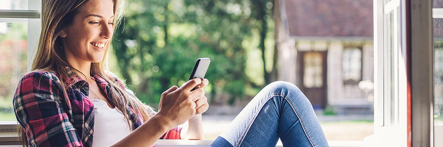 Junge Frau sitzt am offenen Fenster in der Sonne, ihr Blick ist auf ihr Smartphone gerichtet, welches sie mit beiden Händen bedient.