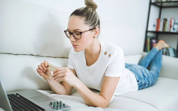 Eine Frau liegt auf einem Sofa und blickt auf den Bildschirm eines Laptops. In ihren Händen hält sie eine Blisterpackung mit kleinen Pillen. Sie wirkt skeptisch.