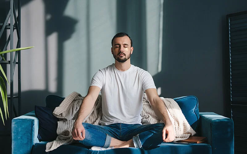 Ein Mann mit geschlossenen Augen sitzt im Schneidersitz auf einem Sofa und legt die Hände auf den Knien ab.