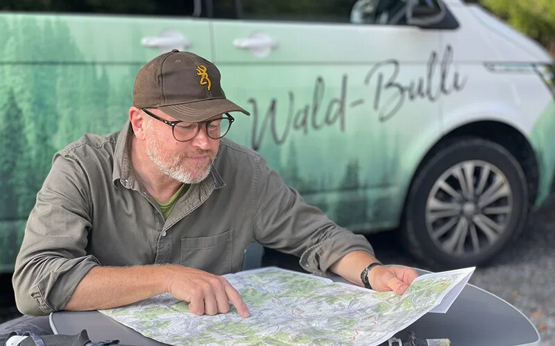 Stefan Pruschwitz sitzt vor einer Landkarte und zeigt darauf, vor seinem Kleinbus im Wald.