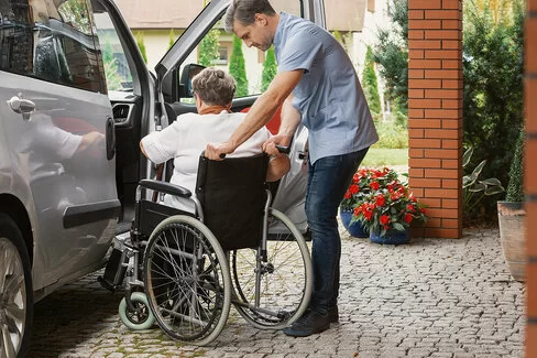 Ein Mann hilft einer Frau im Rollstuhl dabei, in ein Auto einzusteigen. Die AOK übernimmt Fahrkosten in unterschiedlichem Umfang. 