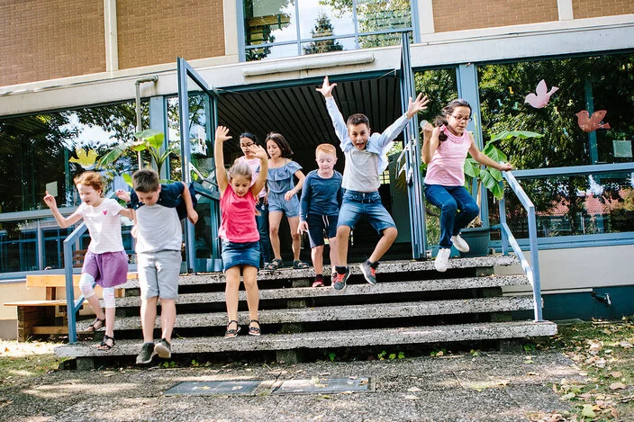 Eine Gruppe Schulkinder springt fröhlich die kurze Treppe am Schuleingang hinunter.