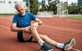 Ein älterer Mann mit Bart in Joggingkleidung sitzt auf einer Laufbahn. Sein rechtes Knie ist angewinkelt. Er fasst sich ans Knie. Sein Gesicht drückt Schmerz aus.