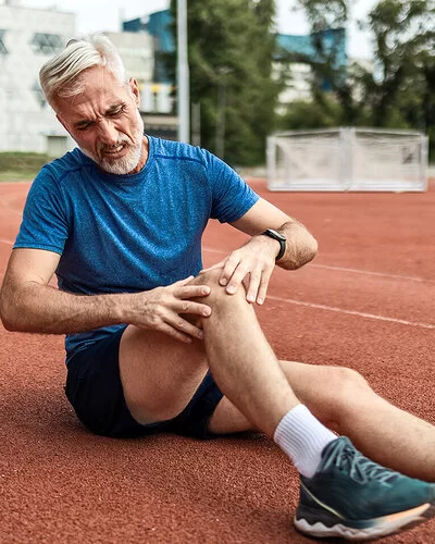 Ein älterer Mann mit Bart in Joggingkleidung sitzt auf einer Laufbahn. Sein rechtes Knie ist angewinkelt. Er fasst sich ans Knie. Sein Gesicht drückt Schmerz aus.